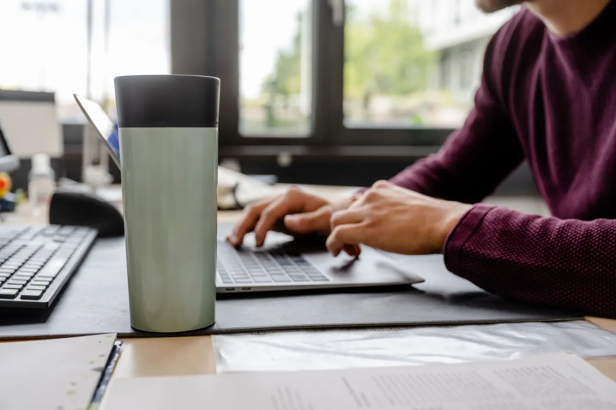 Das Bild zeigt eine Person, die an einem Schreibtisch sitzt und auf einer Tastatur tippt, im Vordergrund ein Kaffeebecher