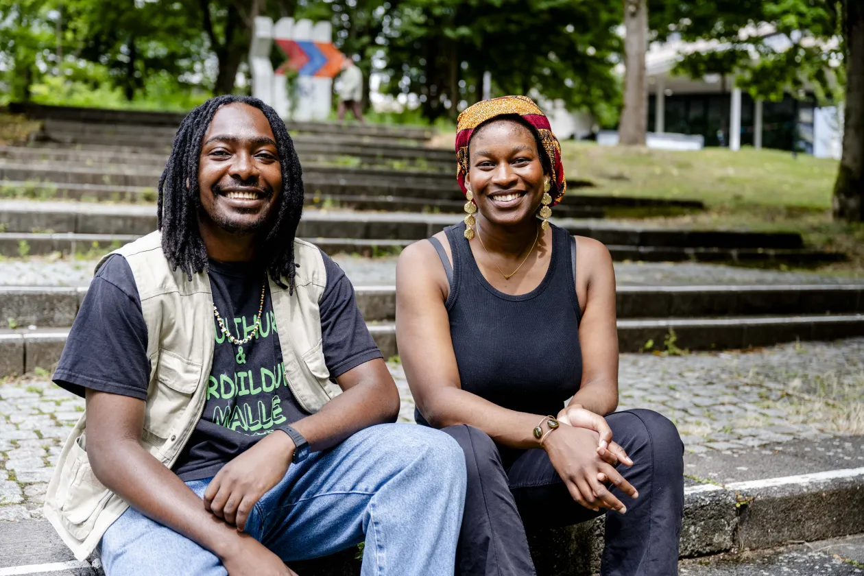 Die Gründer des Start-ups Bissap: ein junger Mann mit Rastalocken sitzt links auf Stufen; eine junge Frau mit buntem Kopftuch sitzt neben ihm; 