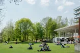 Foto: Eine grüne Wiese, grüne Bäume, ein blauer Himmel und Personen, die auf der Wiese sitzen und liegen.
