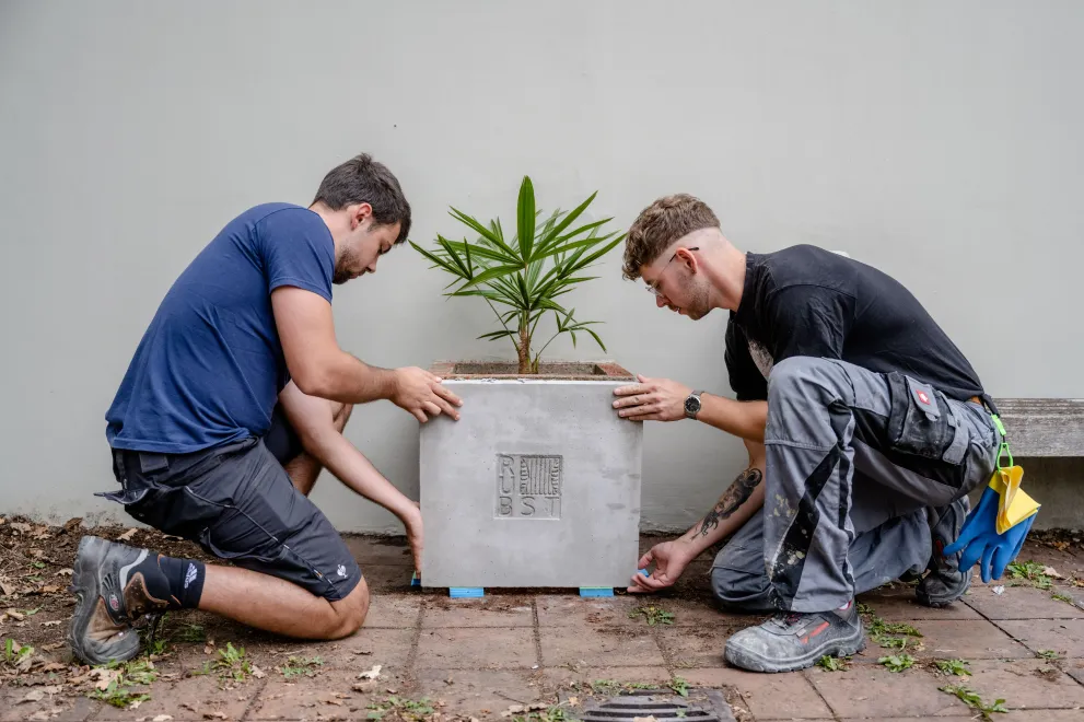 Zwei Männer knien rechts und links eines Betonblumenkastens
