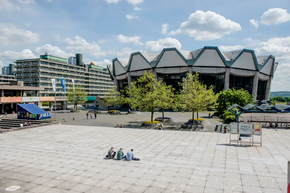 Foto: Foto auf das Forum, das Audimax und das Gebäude NA. Es sitzen wenige Personen auf dem Forumsplatz.