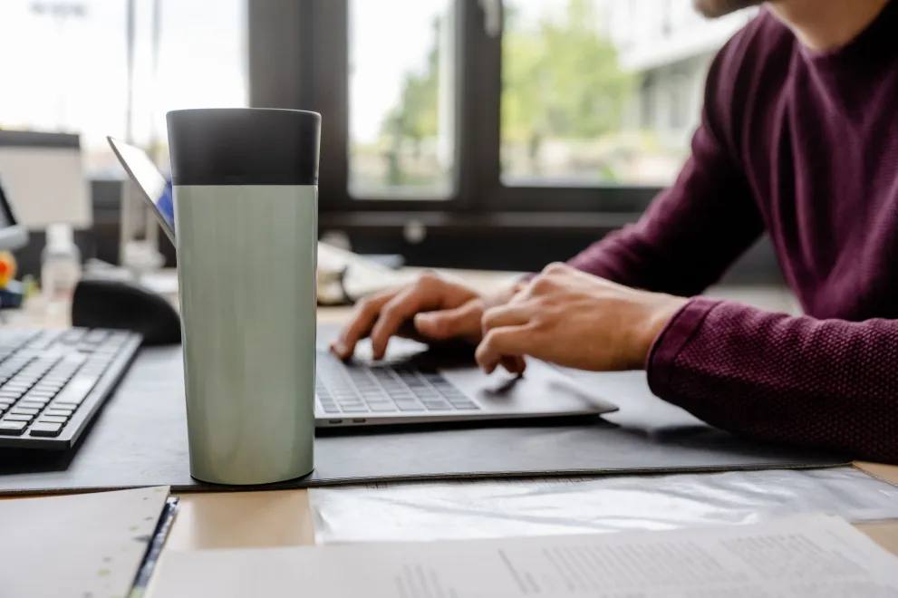 Das Bild zeigt eine Person, die an einem Schreibtisch sitzt und auf einer Tastatur tippt, im Vordergrund ein Kaffeebecher